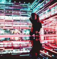 Image of a man kneeling inside a building, with neon lights and reflections, to suggest many dimensions of AI. Image by Drew Dizzy Graham, Unsplash, https://unsplash.com/license